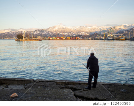 Catcher on spinning in the port area against the backdrop of the mountains. Fishing in Batumi. Lure fishing. Silhouette of a fisherman. Spinning. 122493596