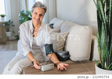 Mature beautiful middle-aged woman with gray hair sits on the sofa and measures blood pressure  122493754