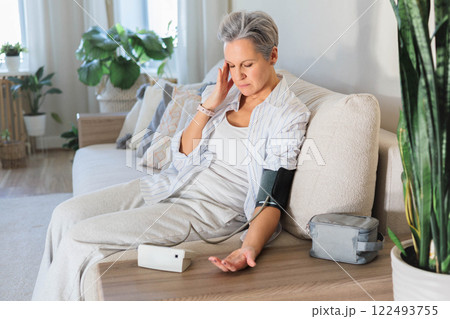 An elderly woman with gray hair sits on the couch and measures blood pressure with a tonometer. An elderly woman with gray hair sits on the couch and measures blood pressure with a tonometer. 122493755