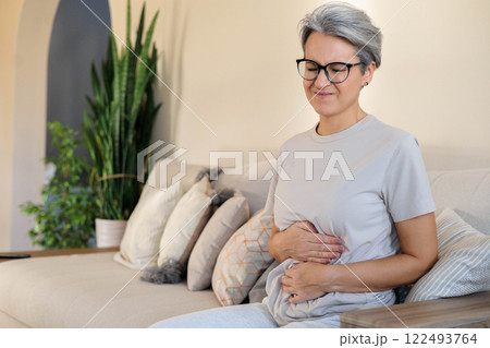 Mature beautiful middle-aged woman with gray hair sits on the couch and holds her stomach  122493764