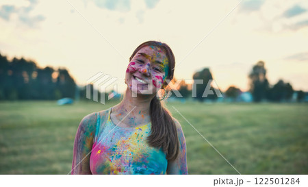 Happy young woman covered in vivid colors, smiling during the holi festival in a field at sunset 122501284