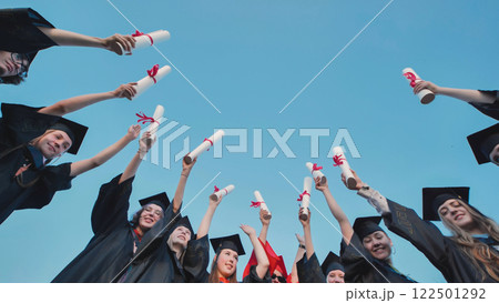 Students in graduation attire celebrating their achievement by raising their diplomas on graduation day Students in graduation attire celebrating their achievement by raising their diplomas on graduation day 122501292