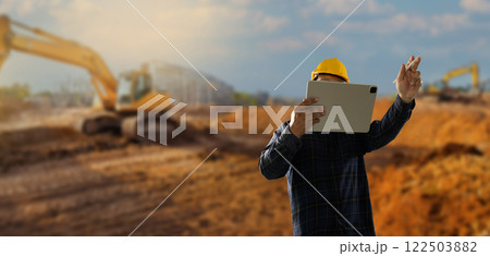 engineer working at site of a large building man wearing a yellow hard hat is looking at a tablet while standing in front 122503882