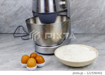 Modern kitchen machine and ingredients for preparing dough (eggs and flour) on a kitchen table Modern kitchen machine and ingredients for preparing dough (eggs and flour) on a kitchen table 122506402