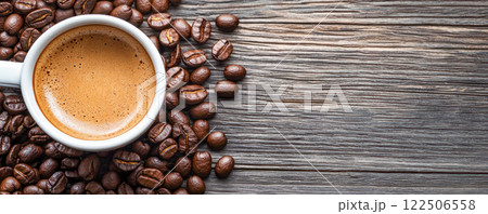 Close-up of a coffee cup with rich crema surrounded by roasted coffee beans on a rustic wooden table. 122506558