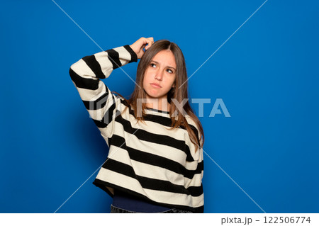Hispanic teenage girl about 15 years old wearing a striped sweater and a puzzled facial expression, scratches her head in frustration, isolated over a blue background. Hmm, I don't remember 122506774