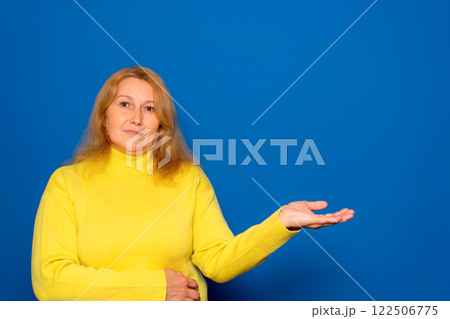 Caucasian woman in her 40s wearing a yellow sweater pointing at something on open palm, demonstrating invisible object, free space for mockup, blue background, panorama. 122506775