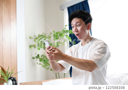 ベッドに座りスマホを持ち驚くアジア人男性 122508108