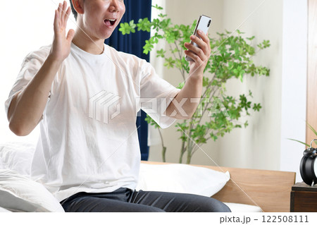 ベッドに座りスマホを持ち驚くアジア人男性 ベッドに座りスマホを持ち驚くアジア人男性 122508111