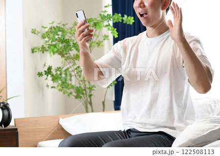 ベッドに座りスマホを持ち驚くアジア人男性 ベッドに座りスマホを持ち驚くアジア人男性 122508113