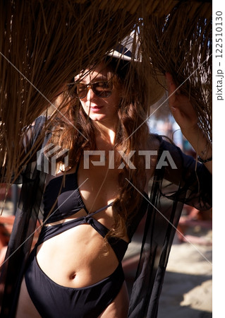 Elegant woman in black bikini under thatched beach umbrella shade  122510130
