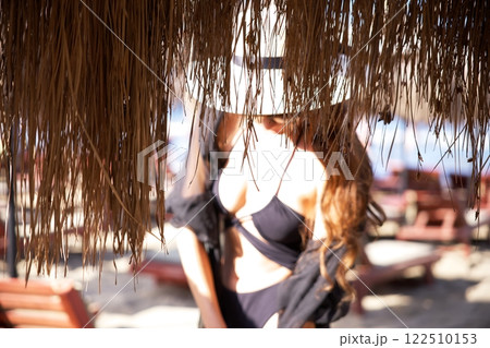 Stylish woman in black bikini under straw beach canopy  122510153
