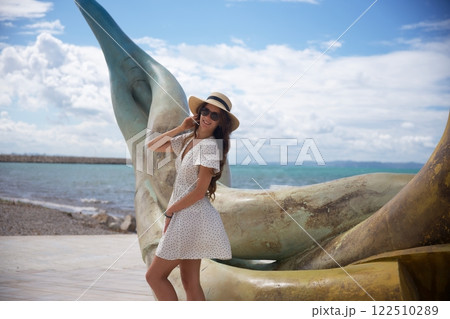 Smiling woman in sun hat posing by seaside sculpture  122510289