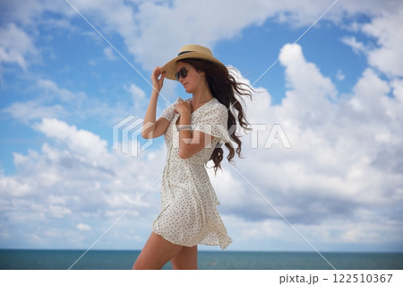 Elegant woman in sun hat adjusting hat in ocean breeze Elegant woman in sun hat adjusting hat in ocean breeze 122510367