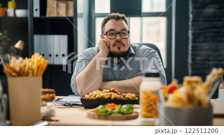 Overweight man sitting at desk with various food items, looking thoughtful and contemplative 122511200