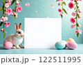 A white square for text in the center of the picture, a light rabbit is sitting next to it and Easter eggs of different colors are lying, pink flowers hanging on top on a blue background. The concept 122511995