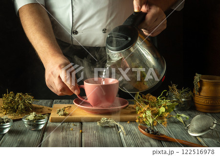 A skilled hand pours steaming water from a kettle into a delicate pink cup, filling it with various herbs laid out on a wooden surface, inviting warmth and comfort into the cozy kitchen atmosphere 122516978