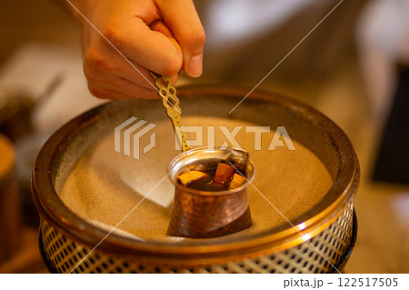 A barista's hand skillfully crafts coffee on sand using a traditional copper Turkish cezve 122517505