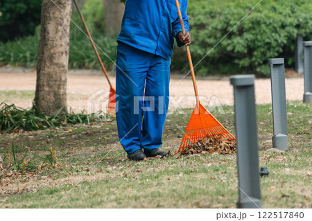 black janitor in blue work clothes, uniform collects withered leaves with rake black janitor in blue work clothes, uniform collects withered leaves with rake 122517840