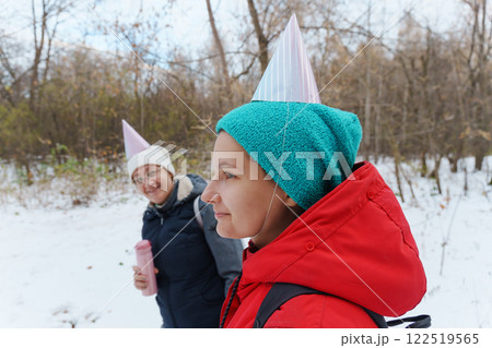 Two friends stroll through a serene, snow-covered forest, donning playful party hats. The atmosphere is cheerful 122519565