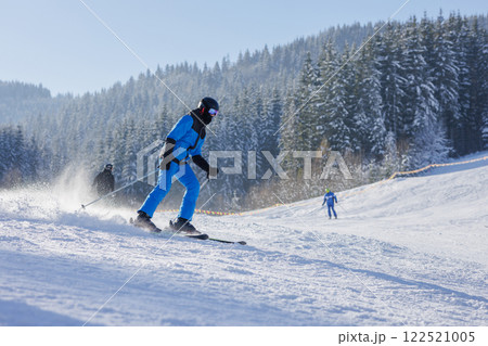 A skier in a blue outfit gracefully descends a snowy slope, surrounded by evergreen trees. The bright winter sun illuminates the scene, creating a picturesque winter landscape. 122521005