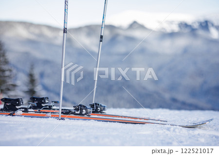 A pair of skis and poles on a snowy mountain slope with a blurred mountain range in the background. 122521017
