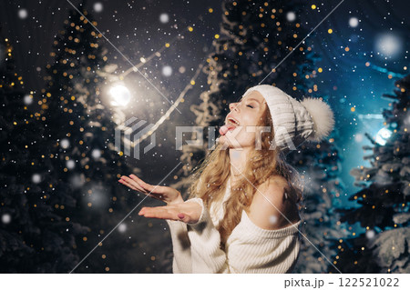 A joyful woman in a cozy sweater and winter hat playfully catches snowflakes with her tongue, in a snowy forest with twinkling lights, a magical winter atmosphere. 122521022