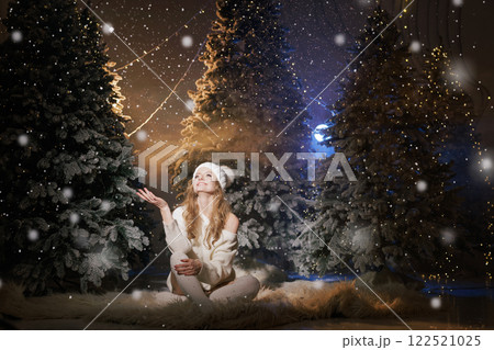A woman in winter clothing sits on a snowy ground surrounded by decorated Christmas trees, with snowflakes falling gently around her. 122521025
