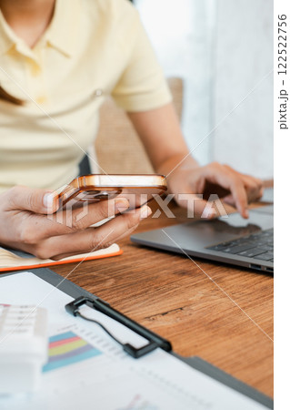 Close-up of a person multitasking with a smartphone and laptop at a desk, surrounded by charts and documents. Close-up of a person multitasking with a smartphone and laptop at a desk, surrounded by charts and documents. 122522756