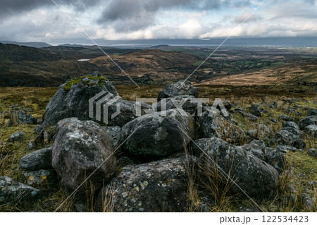 Grey boulders, some moss-covered, are scattered across a hillside. The background shows a vast, rolling landscape under a cloudy sky. Grey boulders, some moss-covered, are scattered across a hillside. The background shows a vast, rolling landscape under a cloudy sky. 122534423