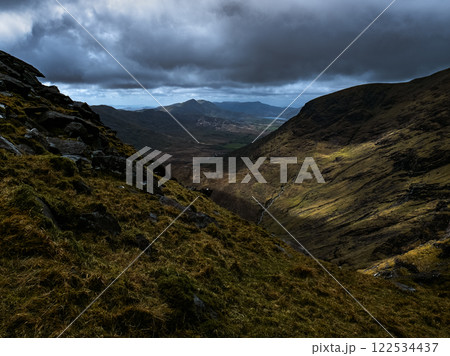 Dark, dramatic clouds fill the upper frame, contrasting with sunlit, rolling green hills in the valley below. A small stream cuts through the center. 122534437