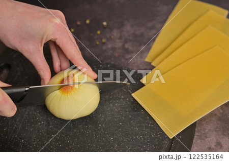 Woman's hand cutting onion on the black kitchen cutting board. Lasagne pasta sheets. Black background. Homemade lasagna cooking preparation Woman's hand cutting onion on the black kitchen cutting board. Lasagne pasta sheets. Black background. Homemade lasagna cooking preparation 122535164