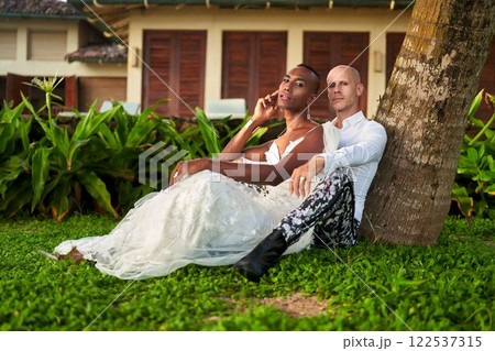 Person in white dress leans on palm tree beside partner in bold outfit. Tropical villa setting highlights lgbtq wedding theme. Inclusive love, diverse celebration, freedom in legal marriage. 122537315