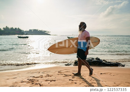 Active senior ethnic man walks on sunny beach with surfboard. Elderly surfer enjoys ocean waves in vibrant lifestyle. Retirement adventure with surf culture. Promoting healthy aging in coastal scene. 122537335