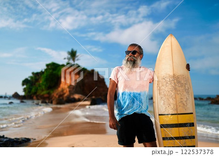Elderly man with surfboard on sunny beach. Active senior enjoying ocean view, healthy retirement lifestyle adventure. Surfing as leisure in golden years. Celebrating vitality, new experiences. 122537378