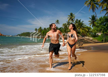 Happy couple runs on tropical beach, hand in hand. Healthy young man, woman in swimwear jog on sandy shore, palm trees in background. Fitness run, sunny travel destination. Active vacation, wellness. 122537398