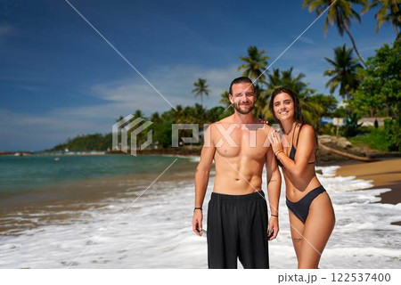 Fit couple stands on tropical beach, man shirtless with toned abs, woman in bikini, both smiling. Active pair enjoys sunny seaside, health, travel lifestyle. Vacation at exotic destination by sea. 122537400