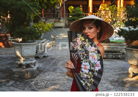 Portrait of a female tourist with a fan in a Vietnamese traditional non la hat at pagoda on a trip to Vietnam in Asia 122538133