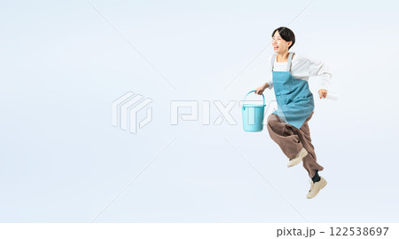 清掃道具を持ってジャンプするエプロンをつけたミドル女性の全身写真 122538697