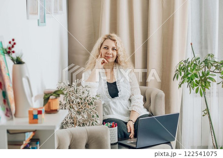 woman with laptop in cozy home office on beige armchair, with plants and in white blouse with curly blonde hair, concept of remote work, business, lifestyle woman with laptop in cozy home office on beige armchair, with plants and in white blouse with curly blonde hair, concept of remote work, business, lifestyle 122541075