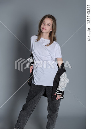 A teenage girl stands confidently in a white t-shirt and jeans, holding a jacket loosely at her side. Her relaxed expression and casual pose reflect a laid-back attitude and effortless style. A teenage girl stands confidently in a white t-shirt and jeans, holding a jacket loosely at her side. Her relaxed expression and casual pose reflect a laid-back attitude and effortless style. 122543843