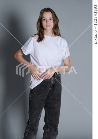 A teenage girl stands confidently in a casual outfit, wearing a white t-shirt and jeans. The simple pose and minimalistic setting reflect a natural and modern sense of style. 122543893