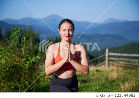 Woman practicing yoga and meditating outdoors in the mountains in serene, natural setting. Portrait of female performing yoga pose, with backdrop of beautiful mountain landscape at sunrise or sunset. Woman practicing yoga and meditating outdoors in the mountains in serene, natural setting. Portrait of female performing yoga pose, with backdrop of beautiful mountain landscape at sunrise or sunset. 122544982