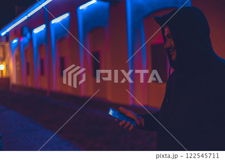 Night city life. A man with a phone in his hand stands at night on a city street illuminated by neon lights. Cyber punk. A man writes a message on his phone on a city street at night. 122545711