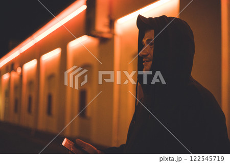 A man in a hood stands on a city street at night with a phone in his hand. The man looks to the side in the light of the city signs at night. Concept of a handsome man on a city street at night. A man in a hood stands on a city street at night with a phone in his hand. The man looks to the side in the light of the city signs at night. Concept of a handsome man on a city street at night. 122545719