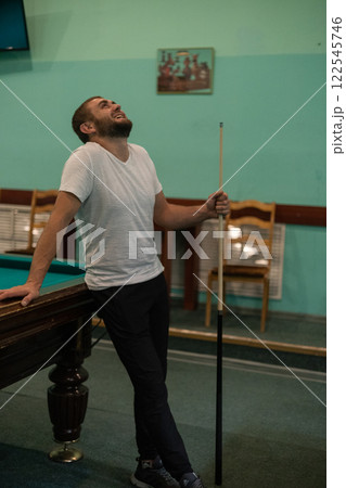 Leaning on the billiard table, the man laughs cheerfully, holding a cue in his hands. A man relaxes and laughs while playing billiards with his friends. Concept of a fun game of billiards. 122545746