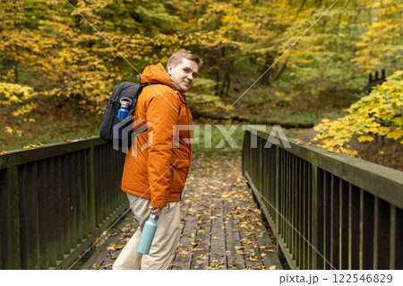 Blonde young man with thermos of tea wearing backpack standing on autumn forest trail, hiking alone. Freedom and active lifestyle concept. High quality photo 122546829