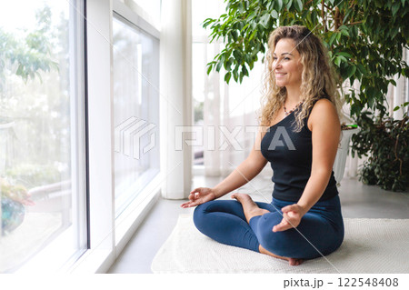 Woman yoga meditation sitting in lotus position near panorama window with houseplant at home Woman yoga meditation sitting in lotus position near panorama window with houseplant at home 122548408