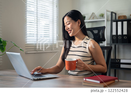 A satisfied Asian businesswoman is enjoying her coffee while working on her laptop in her office. A satisfied Asian businesswoman is enjoying her coffee while working on her laptop in her office. 122552428