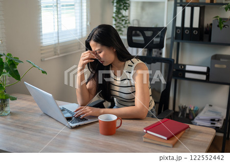 A stressed, tired Asian businesswoman is experiencing severe eye strain while working on her laptop. 122552442